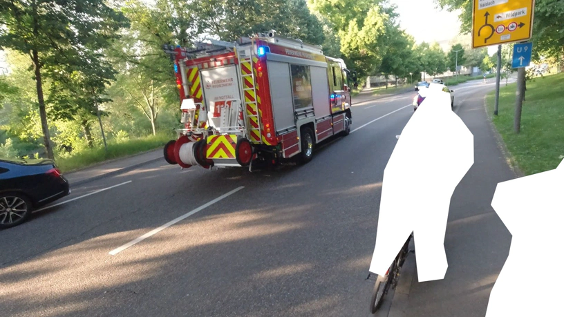 Feuerwehrfahrzeug mittig auf der Straße, links Teil eines stehenden Autos, rechts unkenntlich gemachte Radfahrende, die warten, eine davon auf dem Gehweg.