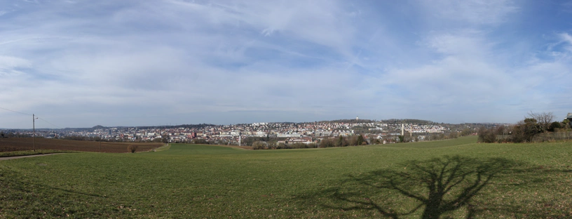 Landschaftspanorama mit blauem, bewölkten Himmel und grünen und braunen Feldern. Im Hintergrund die Stadt Pforzheim.