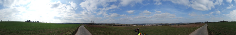 Panorama von einer Landschaft. In der Mitte steht ein Fahrrad. Wolken am Himmel. Grüne Felder.